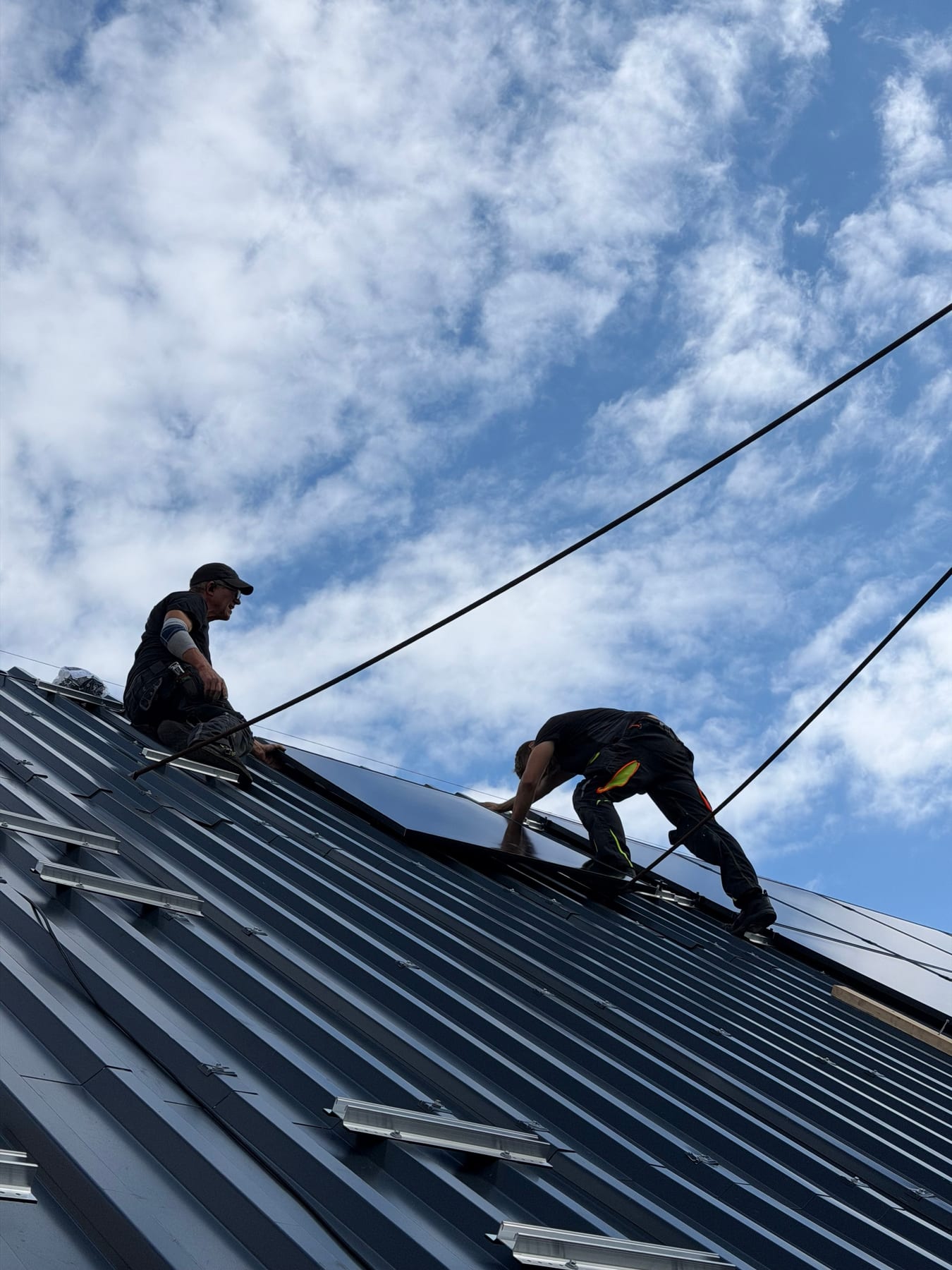 Zwei Monteure auf dem neuen Metalldach beim Verlegen eines Photovoltaikmoduls, gegen den Himmel fotografiert